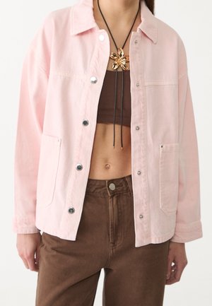 Person trägt eine hellrosa Jacke mit Knöpfen über einem braunen bauchfreien Top und braunen Hosen, ergänzt mit einer goldfarbenen Blumenanhänger-Halskette an einer schwarzen Schnur.