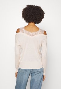 Jasnoróżowy sweter z odsłoniętymi ramionami, zdobiony koronką wzdłuż dekoltu i wykończony prążkowanym obszyciem. Miękka w dotyku tkanina i luźny krój. Styl casualowy.