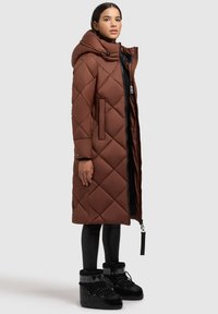 Brūns pārklāts pūkainais mētelis ar kapuci, ar sānu kabatām un kontrastējošu melnu oderi, valkāts virs melnajām legingām un melnajām ziemas zābakiem.