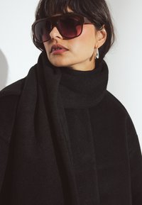 Czarna szalik wykonany z miękkiej, teksturowanej tkaniny, zarzucony na szyję; duże brązowe okulary przeciwsłoneczne z gradientowymi szkłami i widocznym wiszącym kolczykiem w kształcie kropli.