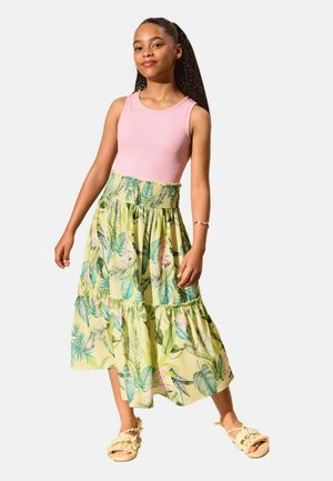 Ein junges Mädchen steht und trägt ein rosa ärmelloses Oberteil, einen grünen geblümten Midi-Rock und geflochtene Sandalen mit Knöchelriemen vor einem schlichten hellen Hintergrund.