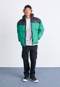 Chaqueta acolchada verde y negra con un alto cuello, llevada sobre una camisa blanca. Pantalones cargo negros y zapatillas negras completan el look.