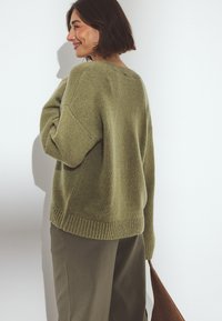 Alyvučių žalios spalvos megztinis su laisvu siluetu, guma aptrauktos rankovės ir nukritusiais pečiais, derinamas su tamsiai žaliais kelnais ir ruda rankine.