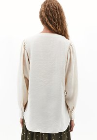 Lichtbeige blouse met lange, puffed mouwen. Heeft een ronde halslijn en knoopmanchetten. Gemaakt van een gestructureerde, lichte stof.