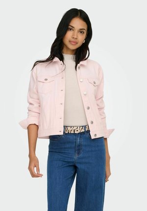 Ung kvinne med langt mørkt hår iført lys rosa jakke med knapper, beige topp, blå jeans og mønstret belte, stående foran hvit bakgrunn.