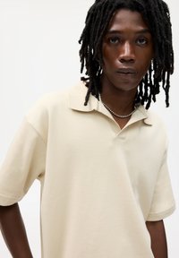 Beige kortærmet polo shirt med krave, der har et tekstureret stof og en afslappet pasform, båret med en sølv kæde. 