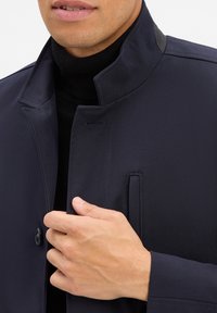 Dunkelblaue Jacke mit hohem Kragen, ausgestattet mit einem Knopfverschluss und seitlichen Taschen, aus glattem Stoff mit strukturiertem Design.