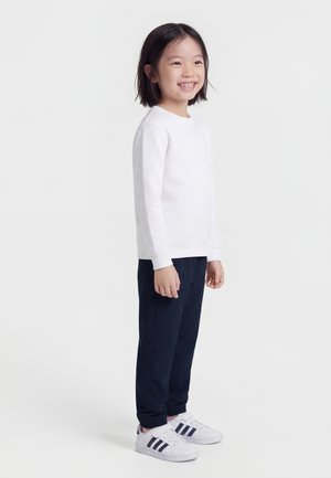 Jeune fille souriante, debout, portant un haut blanc à manches longues, un pantalon bleu marine et des baskets blanches avec des rayures foncées sur un fond blanc uni.