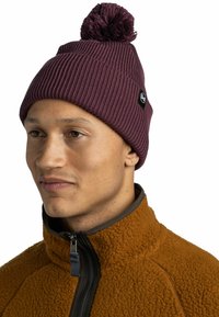 Maroon ribbed beanie with a pom-pom on top and a black logo tag. Worn with a brown fleece jacket featuring a zippered collar.