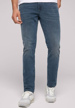 Slim-fit blue denim jeans with a classic five-pocket design and subtle whiskering details, paired with white athletic shoes.