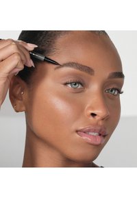 Ołówek do brwi w kolorze czarnym trzymany w ręce obok wyraźnie zdefiniowanych brwi. Osoba ma gładką, jednolitą cerę i błyszczące, neutralno-tonowe usta.