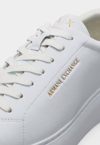 Baskets en cuir blanc avec des lacets blancs, texte doré "ARMANI EXCHANGE" sur le côté, et petit logo doré "AX" sur la languette.