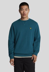 Sudadera verde azulado con cuello redondo y puños acanalados. Presenta un pequeño logotipo amarillo en el lado izquierdo del pecho. Hecha de una tela suave y texturizada.