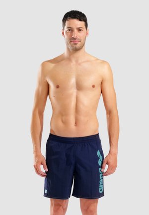 Skjorteløs mand med kort mørkt hår iført marineblå badeshorts med turkis "arena"-logo, der står mod en ensfarvet lys baggrund.