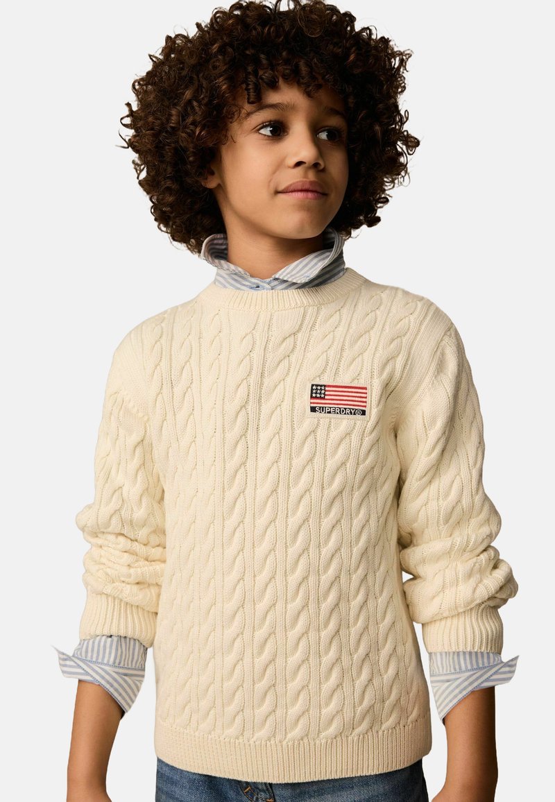 Beige Pullover aus Kabelstrick mit gerippten Bündchen und Saum, versehen mit einem kleinen Aufnäher mit einer amerikanischen Flagge und dem Markenlogo. Darunter ein hellblaues gestreiftes Hemd.
