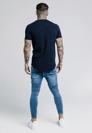 Mężczyzna z tatuażami na szyi i ramionach, ubrany w ciemny, kropkowany t-shirt, obcisłe niebieskie dżinsy i białe trampki, stojący tyłem.