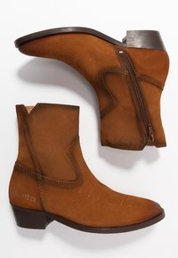 Botines de ante marrón con punta afilada, cremallera lateral y costuras decorativas, con un tacón bajo y una suela de cuero suave.