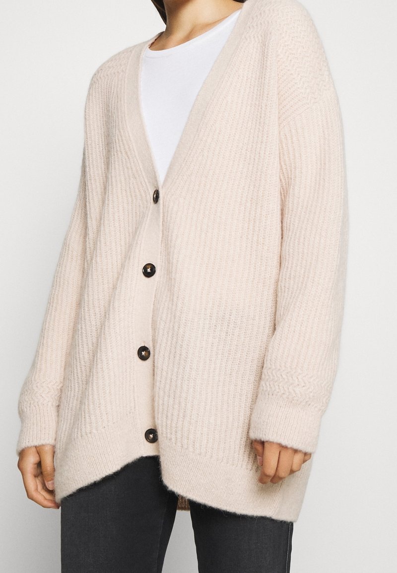 Femme portant un cardigan tricoté côtelé de couleur crème avec de grands boutons foncés par-dessus une chemise blanche et un pantalon noir.