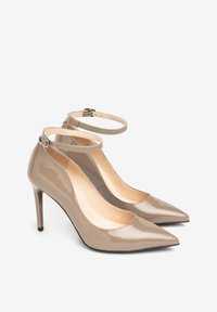 Pár lesklých béžových lodiček na vysokém podpatku se špičatými špičkami a kotníkovými pásky se systémem malých přezek na bílém pozadí.