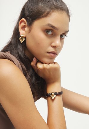 Mujer joven con cabello largo y oscuro, apoyando la barbilla en el puño, lleva pendientes dorados en forma de corazón y una pulsera a juego con cordón marrón.