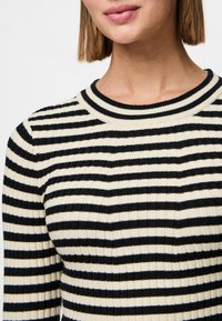 Femme portant un pull côtelé à rayures horizontales noires et blanches avec un col rond, visible des épaules jusqu'à la bouche.