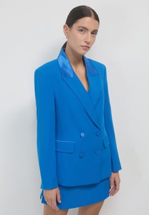 Blazer blu a doppio petto con lapels in satin, due tasche frontali e chiusure a bottone. Abbinato a una gonna corta coordinata. Texture liscia.
