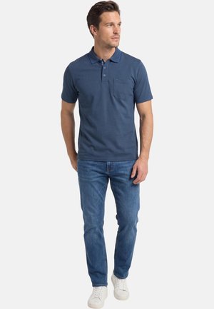 Homme portant un polo manches courtes bleu marine, un jean bleu et des baskets blanches, debout et regardant sur le côté.