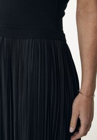 Černá skladaná sukně z texturované látky. Vrchní část má žebrovaný pas. Náramek zdobí ruku odloženou na sukni.