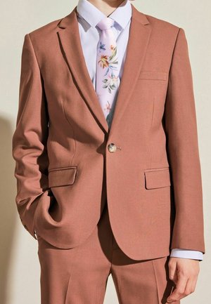 Homme portant un costume couleur rouille avec une chemise blanche et une cravate rose pâle à motif floral, main dans la poche, debout devant un fond uni.