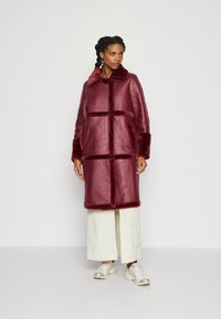 Bordeaux faux fur frakke med krave, store lommer og farveblokering design. Mellemlang, glat tekstur og løs pasform.