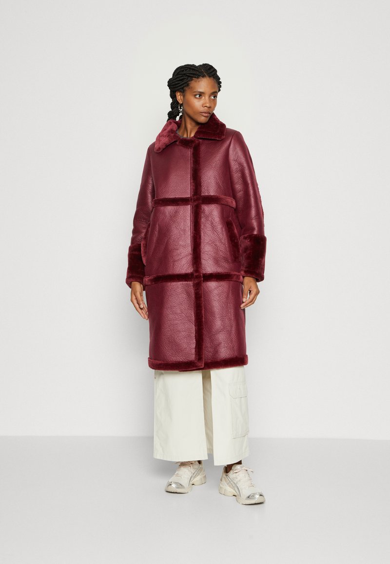 Bordeaux faux fur frakke med krave, store lommer og farveblokering design. Mellemlang, glat tekstur og løs pasform.