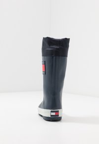 Tmavě modrá gumová dešťová bota s texturovaným černým okrajem, bílou podrážkou a červenobílým logem na straně. Vysoký, štíhlý design; hladký povrch.