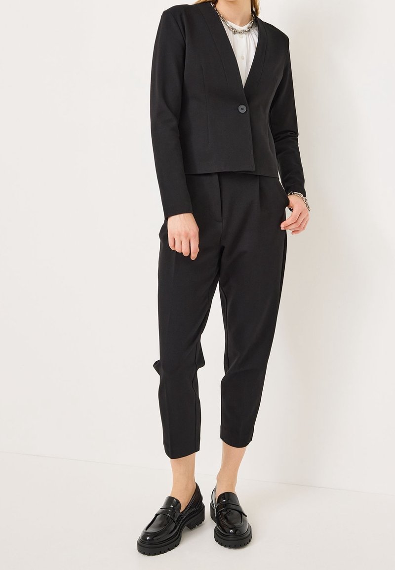 Blazer noir ajusté avec un col en V, fermeture à un bouton, accompagné d'un pantalon court assorti et de mocassins noirs élégants.