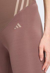 Leggings d'une couleur mauve douce, fabriqués à partir d'un tissu lisse et extensible. Présentent trois rayures beiges et un petit logo de marque sur la cuisse.