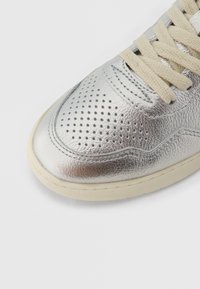 Hopeanhohtoinen hopeinen nahkatennari rei'itetyllä kuviolla, kermanväriset nauhat ja kumipohja. Ominaisuuksiin kuuluu sileä pinta ja pyöristetty kärki.