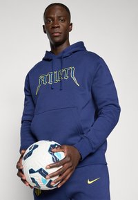 Ung mand i blå hættetrøje og bukser, der holder en hvid og blå fodbold foran en ensfarvet grå baggrund.