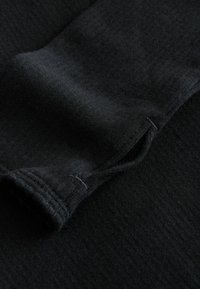 Schwarzer Stoff mit gerippter Textur, der eine Nahaufnahme eines Ärmelbündchens und eines Nahtdetails zeigt und ein weiches, dehnbares Material präsentiert.