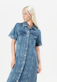 Abito midi in denim con maniche corte, colore azzurro chiaro, chiusura frontale a bottoni, due tasche sul petto e dettagli di cuciture verticali.