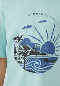 Hellblaues T-Shirt mit einem Grafikmotiv von Palmen, Wellen und Sonne, ergänzt durch den Text "DER SOMMER RUFT" in einem kreisförmigen Design.