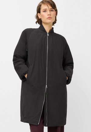 Woman wearing a long black zip-up coat with hands in pockets, standing against a plain light background.