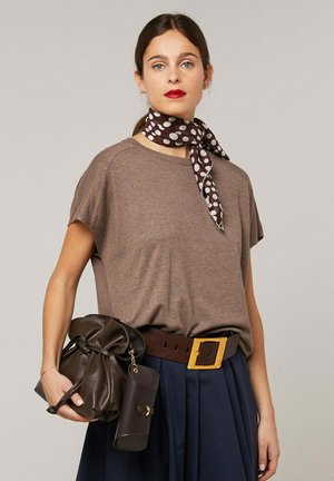 Frau trägt braunes kurzärmliges Oberteil, marineblauen plissee Rock, breiten braunen Gürtel mit großer Schnalle, gepunktetes Halstuch und hält eine dunkelbraune Ledertasche.