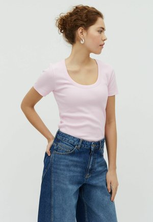 Młoda kobieta z kręconymi włosami związanymi w kok, ubrana w jasnoróżowy t-shirt z dekoltem w serek i wysokim stanem szerokie dżinsy, stoi z jedną ręką na biodrze.