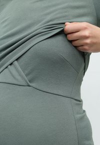 Άτομο που σηκώνει ένα γκριζοπράσινο υφή υφάσματος, δείχνοντας μια καμπυλωτή ραφή σε ένα στενά εφαρμοστό ένδυμα.