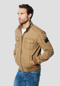 Béžová bomber bunda se dvěma kapsami na hrudi a žebrovanými manžetami. Vyrobena z pevné látky, má vysoký límec a texturovanou nášivku na rukávu.