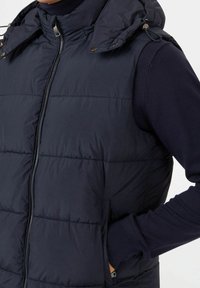 Chaleco acolchado azul marino con cremallera frontal, capucha ajustable y bolsillos laterales. Cuenta con un patrón acolchado y puños acanalados para un ajuste mejorado.