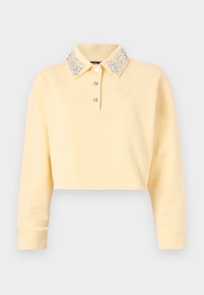 Rövidített sárga pulóver hosszú ujjal, díszített gombos nyakkal, ezüst gyöngyökkel, három gomb a frontra, és hímzett logo.