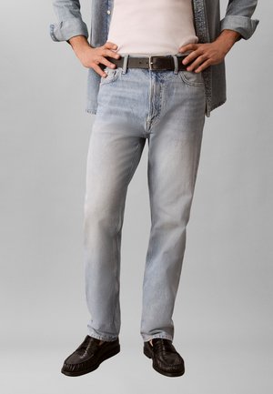 Άτομο που φοράει ανοιχτό μπλε τζιν, μαύρη ζώνη, μαύρα μοκασίνια, λευκό πουκάμισο και ανοιχτό τζιν μπουφάν, στέκεται με τα χέρια στη μέση.