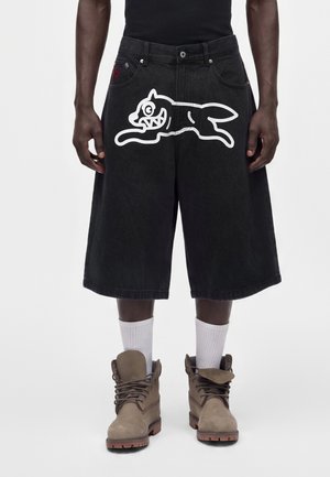 Sorte lange shorts med et hvidt tegneseriehunddesign foran, båret med hvide sokker og beige snørestøvler.