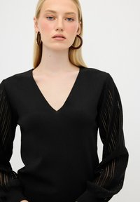 Schwarzer Strickpullover mit tiefem V-Ausschnitt und strukturierten, transparenten langen Ärmeln mit vertikalen Streifen und gerippten Bündchen.