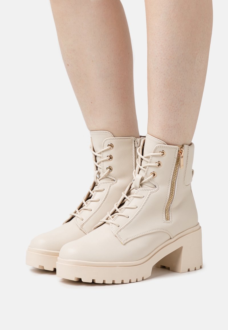 Bottes beiges à lacets montant à la cheville, à talons épais et fermetures éclair latérales, portées sur des jambes nues sur un fond blanc.
